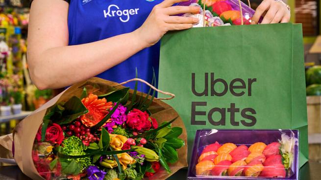 A person wearing a Kroger uniform places sushi into an Uber Eats bag. Flowers sit on a table.